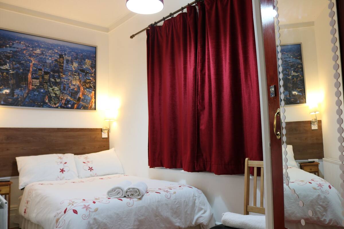 Ein Hotelzimmer mit zwei Betten, roten Vorhängen, einem Stadtbildgemälde und einem Spiegel mit dekorativem Rahmen.