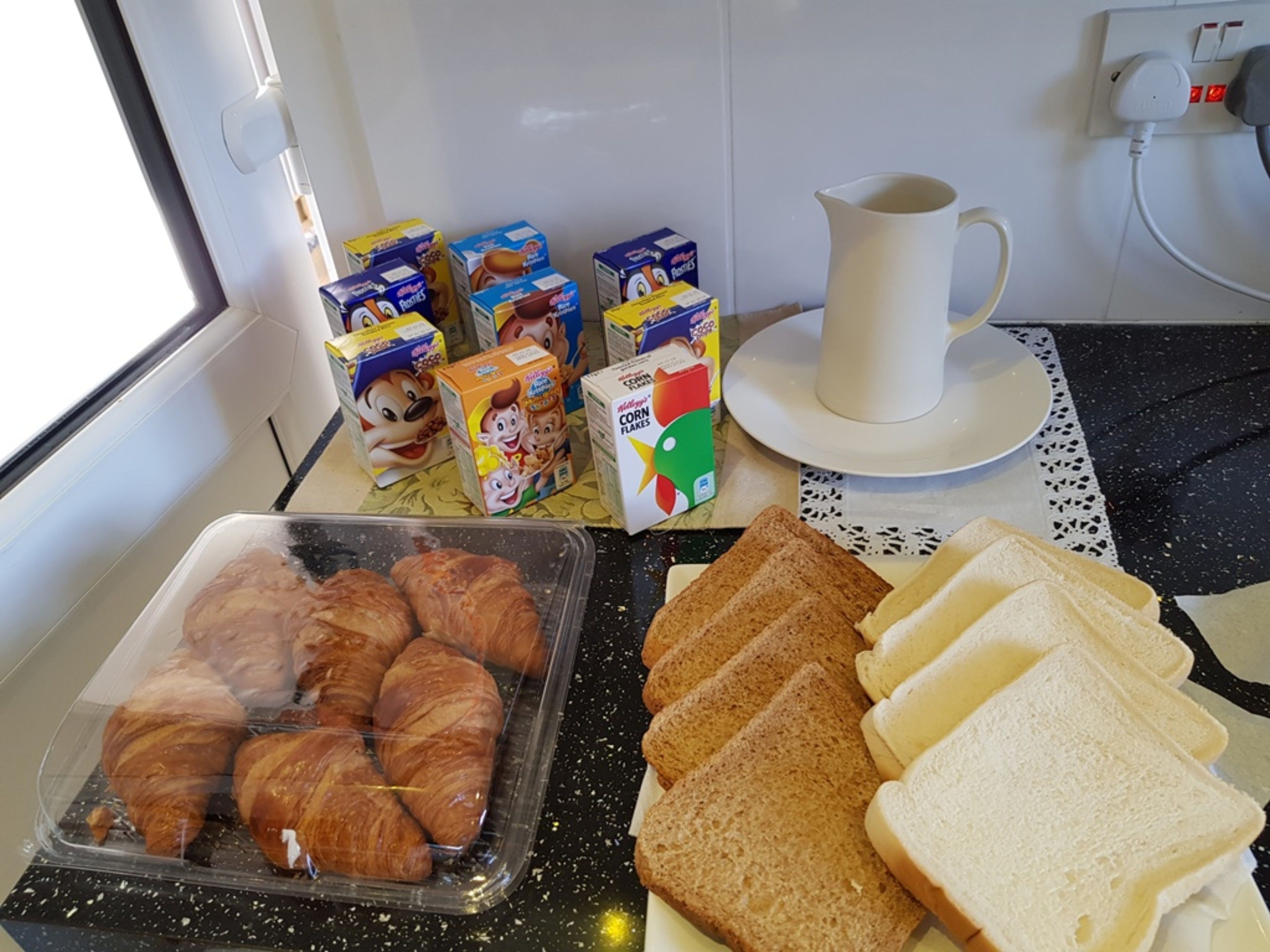 A breakfast setup with croissants, bread, cereal boxes, and a coffee cup on a kitchen counter.