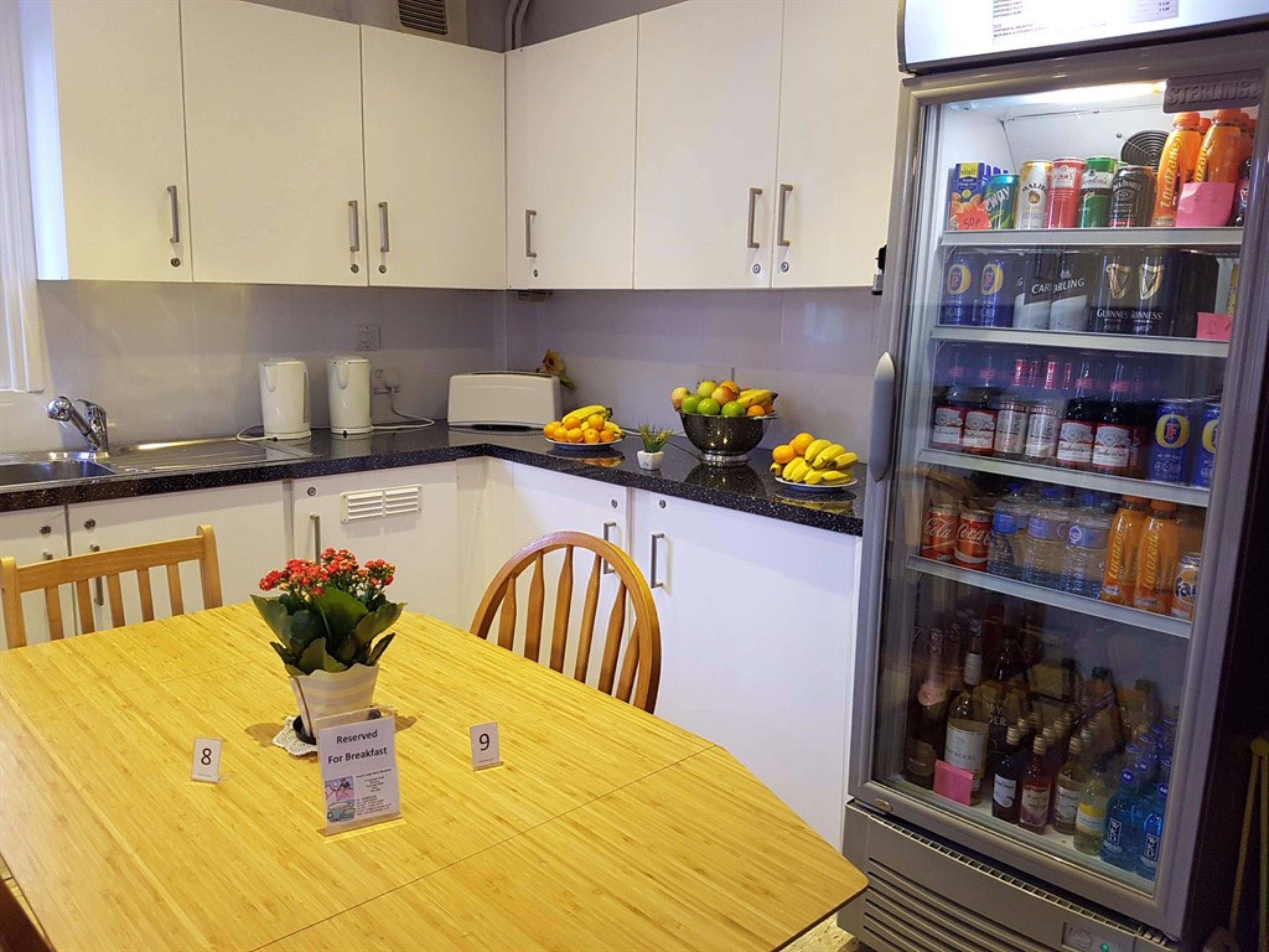 A kitchen with white cabinets, a refrigerator stocked with beverages, a fruit bowl, and a table with breakfast reservation signs.