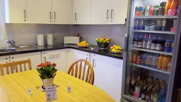 A kitchen with white cabinets, a refrigerator stocked with beverages, a fruit bowl, and a table with breakfast reservation signs.