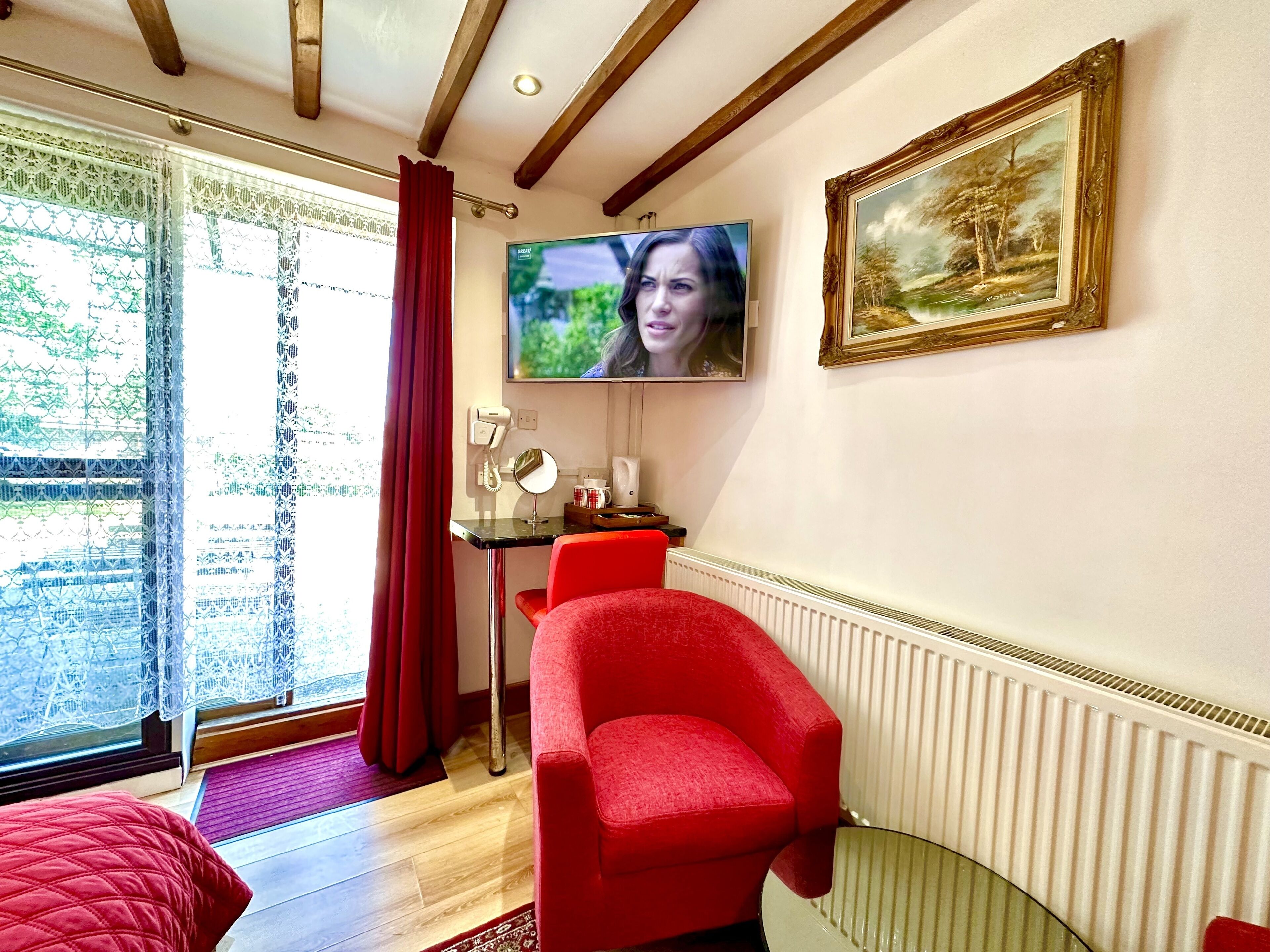 Une pièce avec un fauteuil rouge, un téléviseur fixé au mur, un tableau encadré et une fenêtre avec des rideaux.