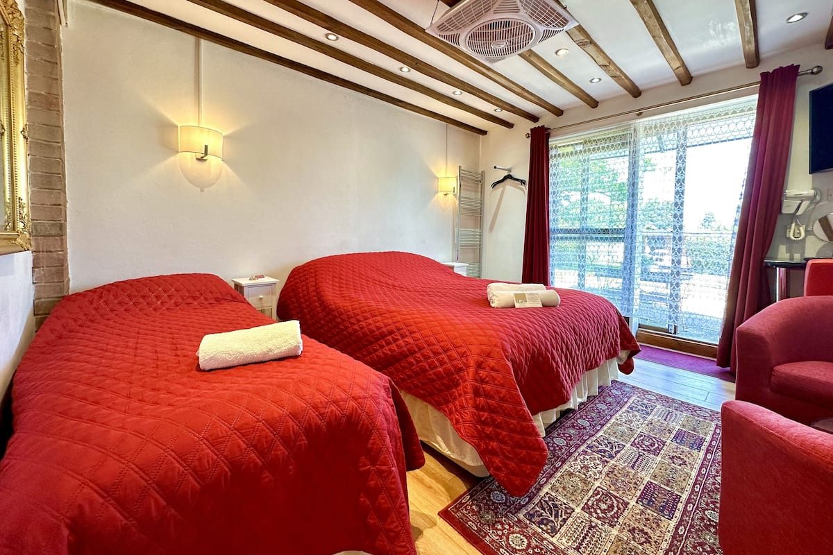 Ein Hotelzimmer mit zwei Betten, einer roten gesteppten Bettdecke, einem gemusterten Teppich, einem Deckenventilator und Blick ins Freie durch ein Fenster mit Vorhang.