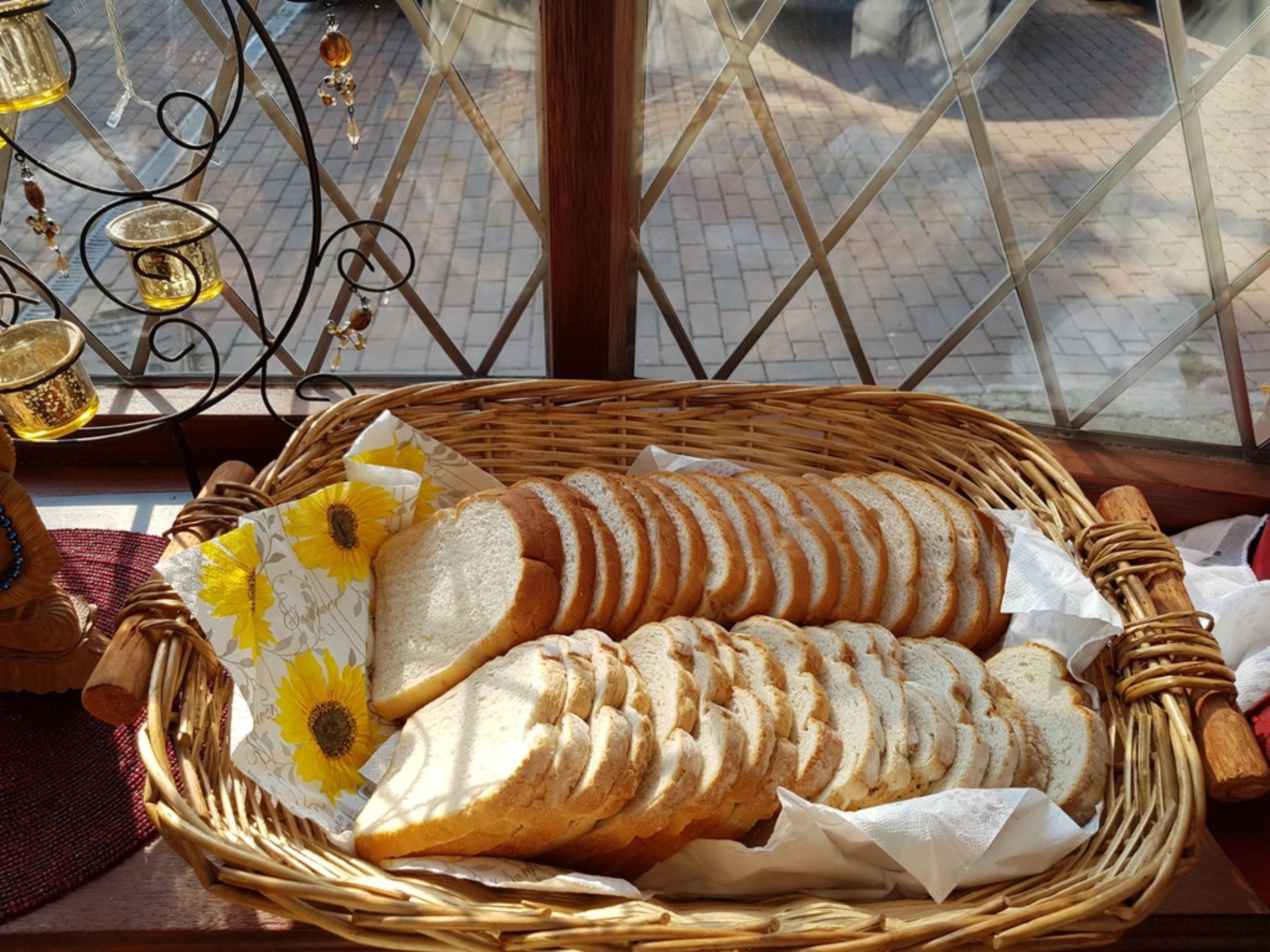 A wicker basket containing sliced bread and a sunflower-patterned napkin, placed on a table near a window with a view of a brick-paved street.