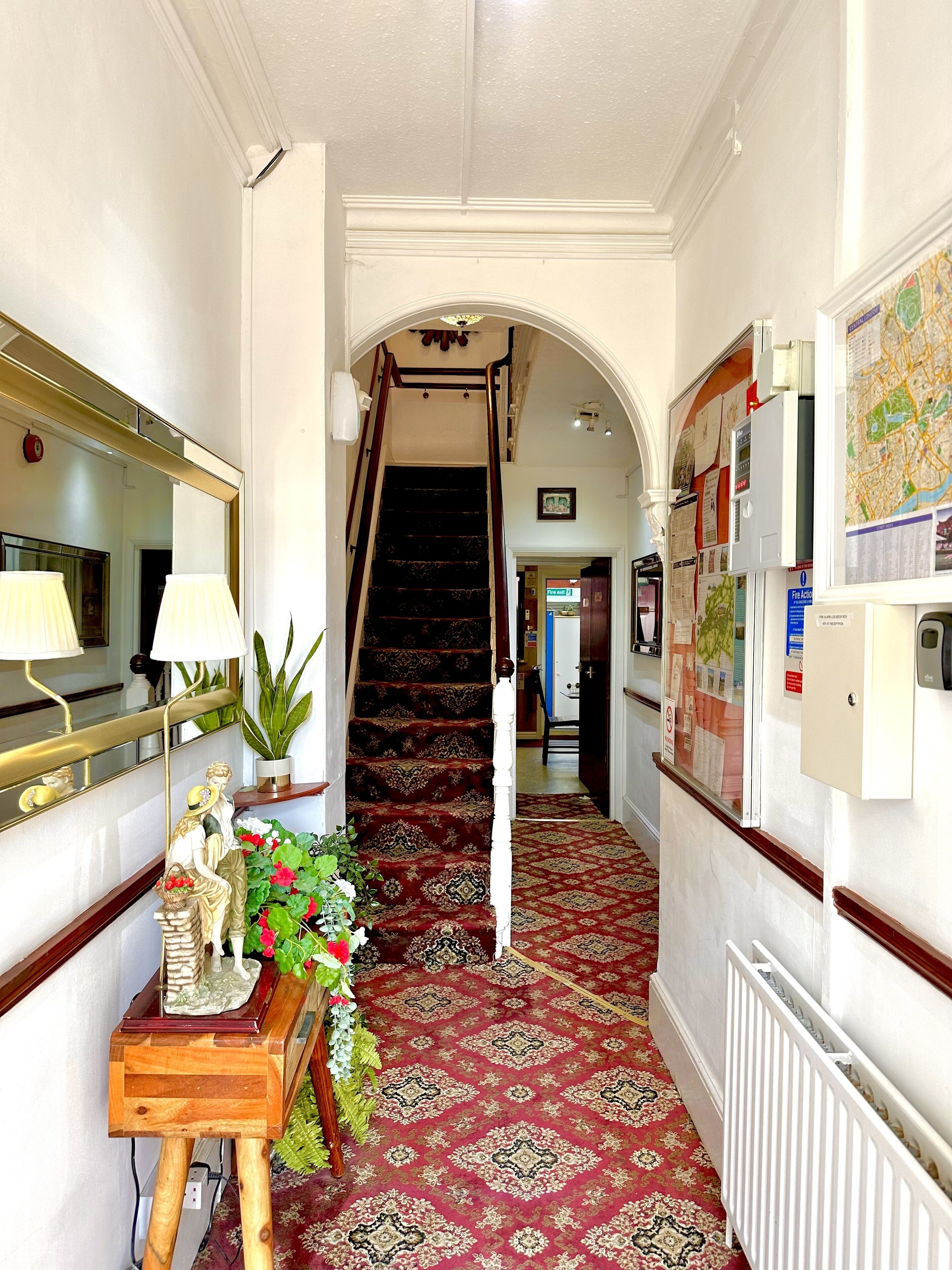 Un couloir avec une moquette rouge, un escalier, un bureau en bois avec une lampe et un miroir.
