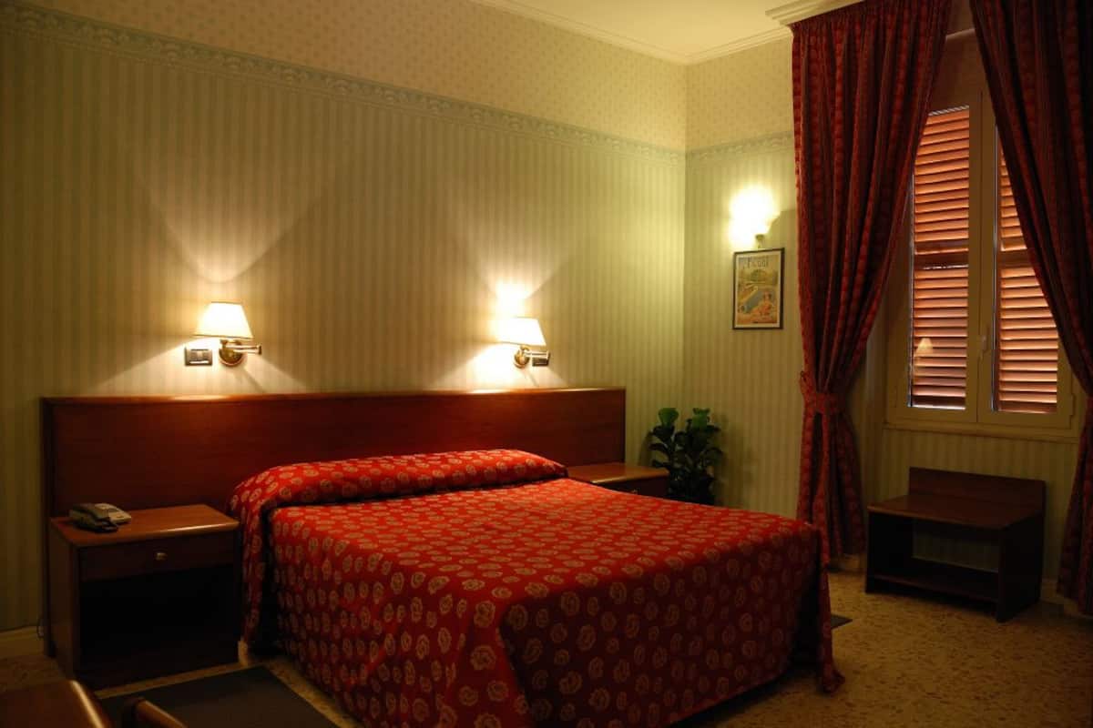 A hotel room with a large bed, two bedside lamps, a nightstand, a small table, and a window with blinds.