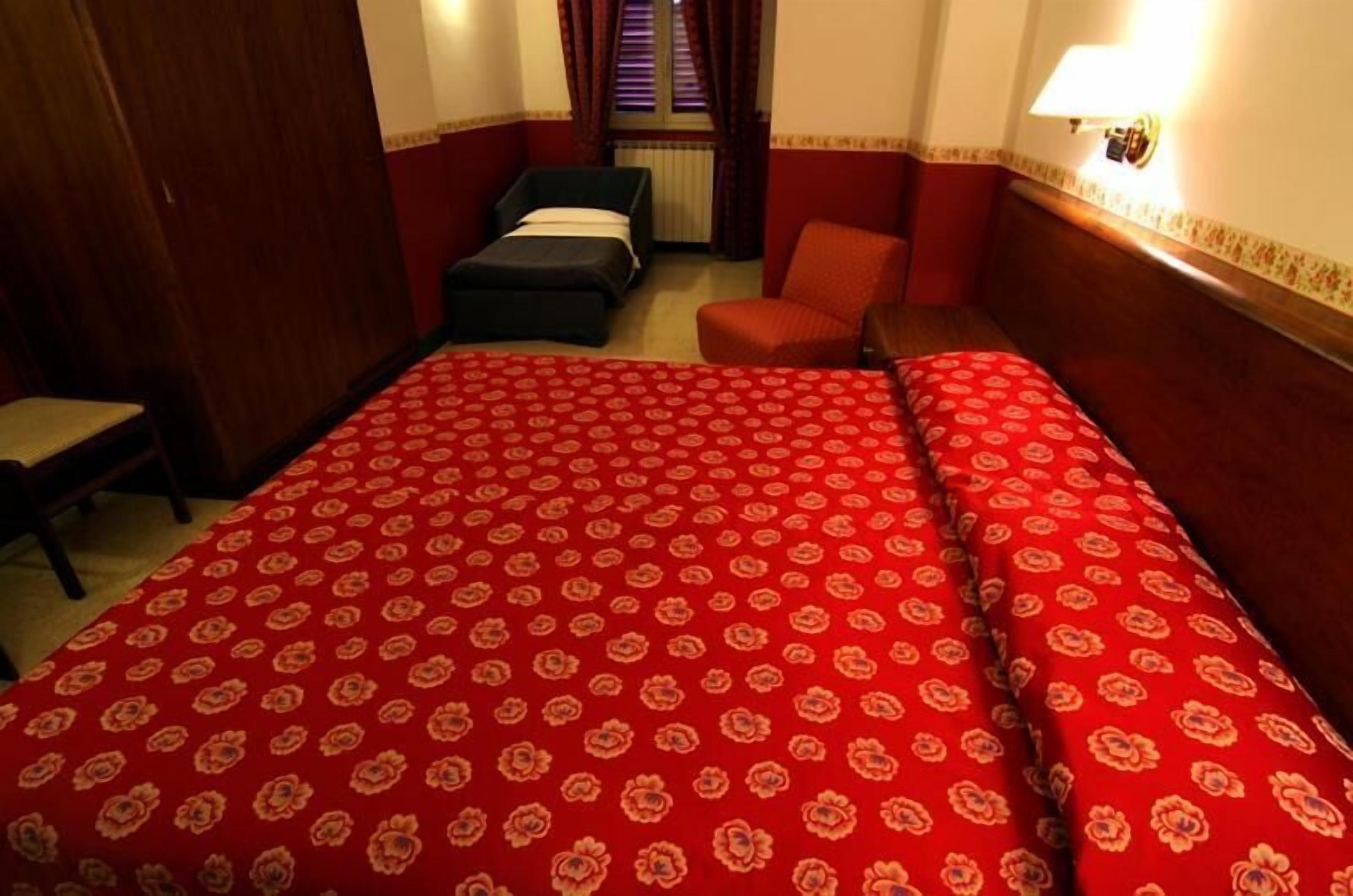 A hotel room with a large bed, a single chair, a small table, and a window with curtains.