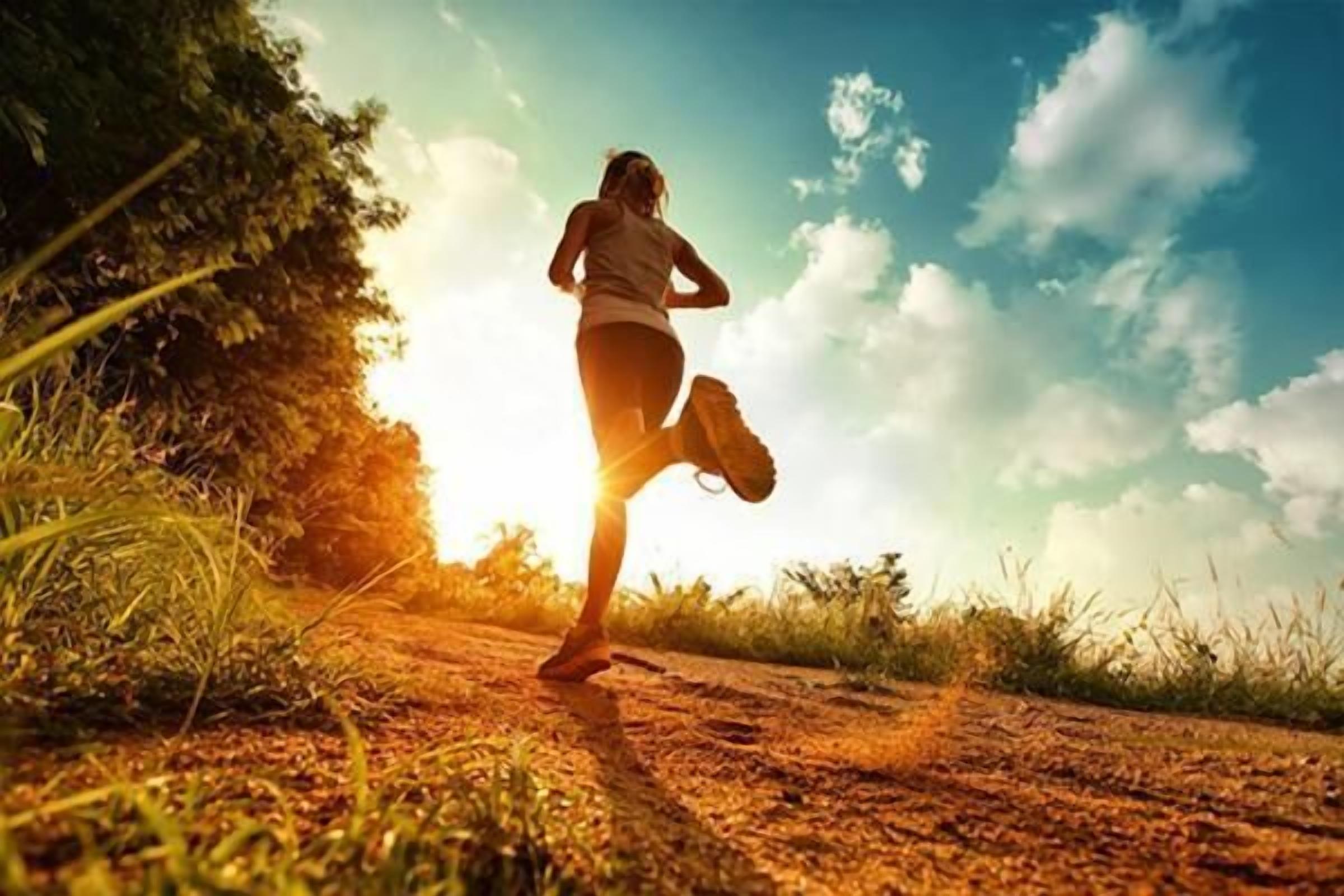 A person running on a dirt path surrounded by grass and trees, with a clear sky in the background.