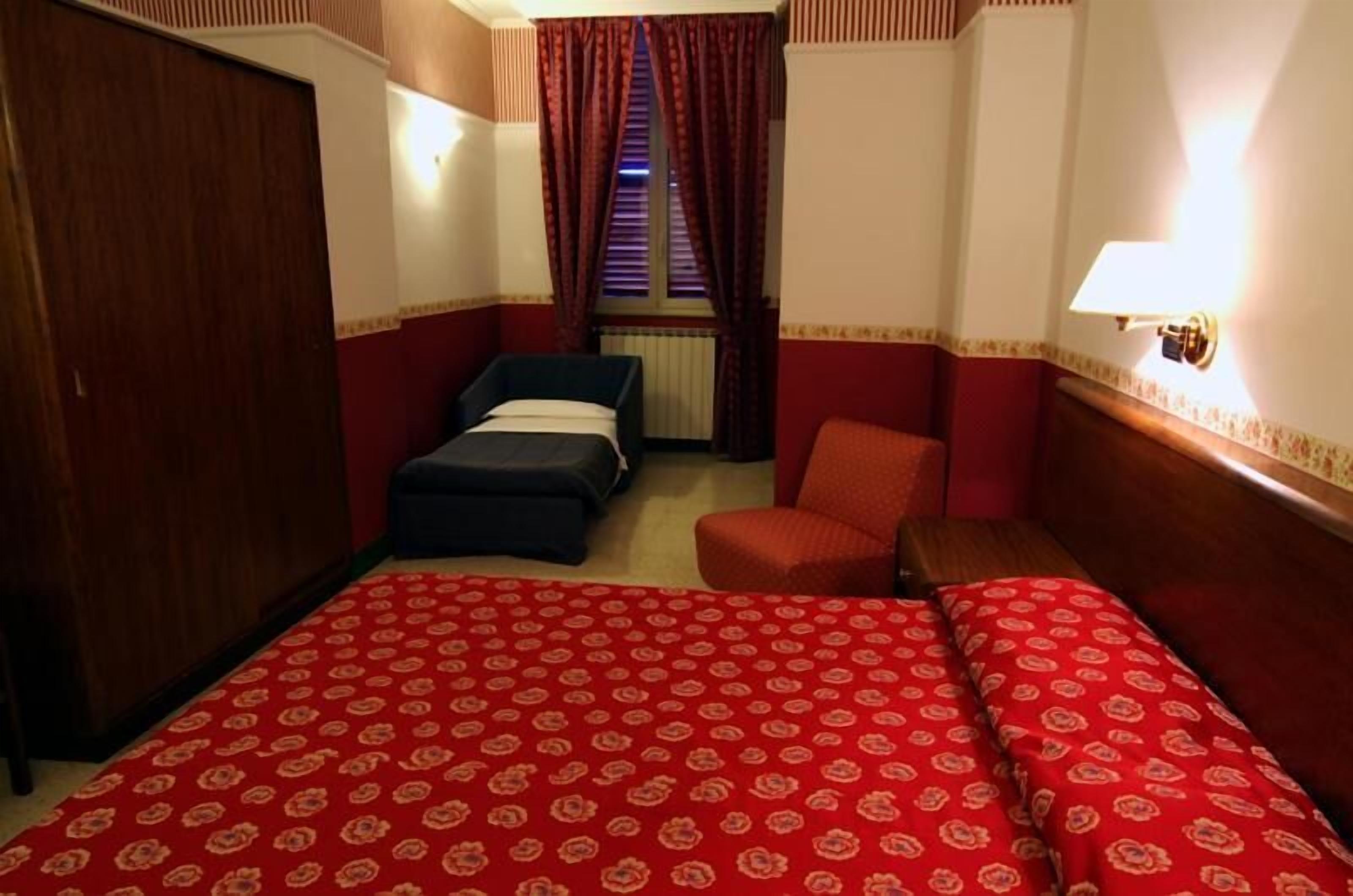 A hotel room with a bed, a chair, a wardrobe, and a window with curtains.