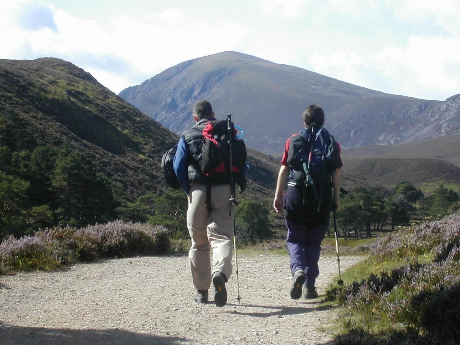 Twee wandelaars op een grindpad, omringd door heuvels en schaarse begroeiing.
