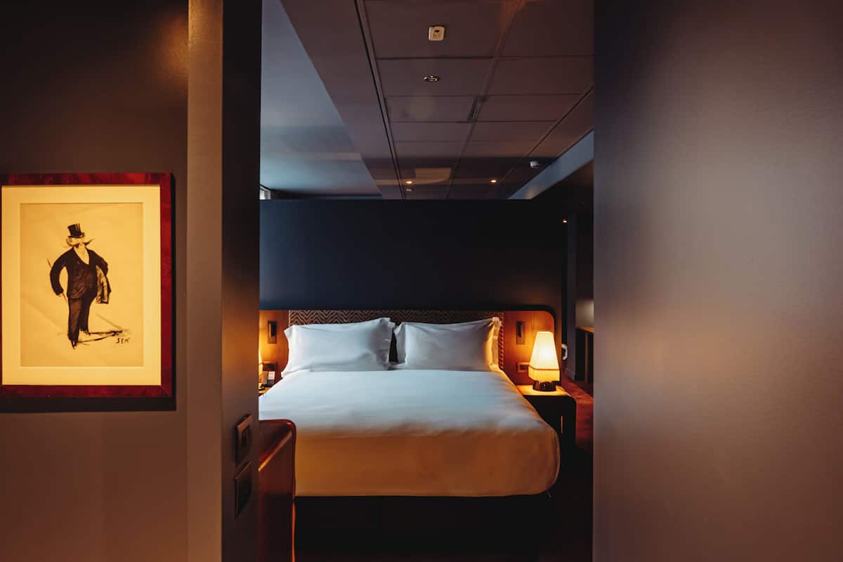 A hotel room with a large bed, a framed artwork on the wall, and a nightstand with a lamp.