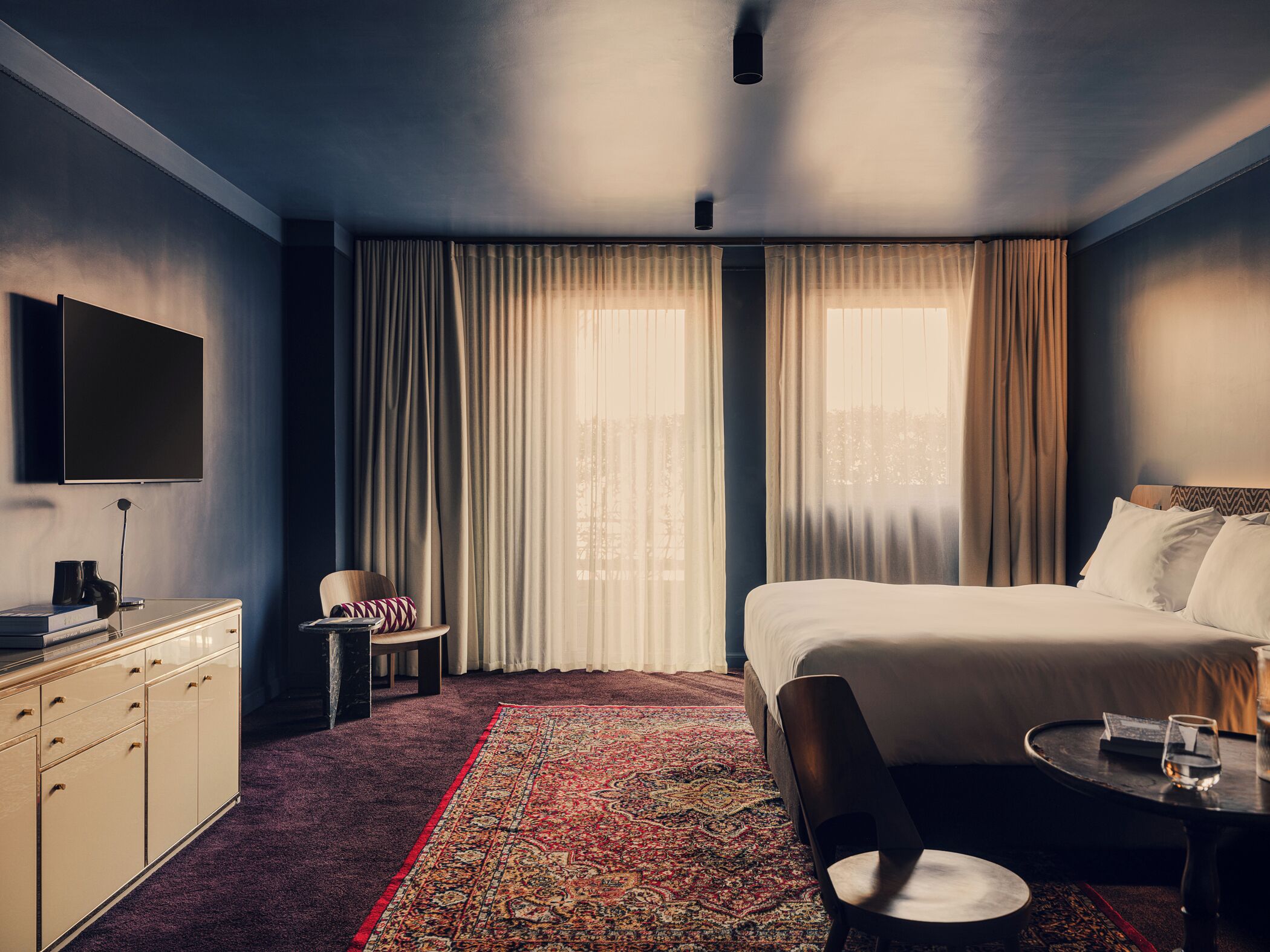 A hotel room with a bed, a desk, a chair, a small table, and a TV mounted on the wall.