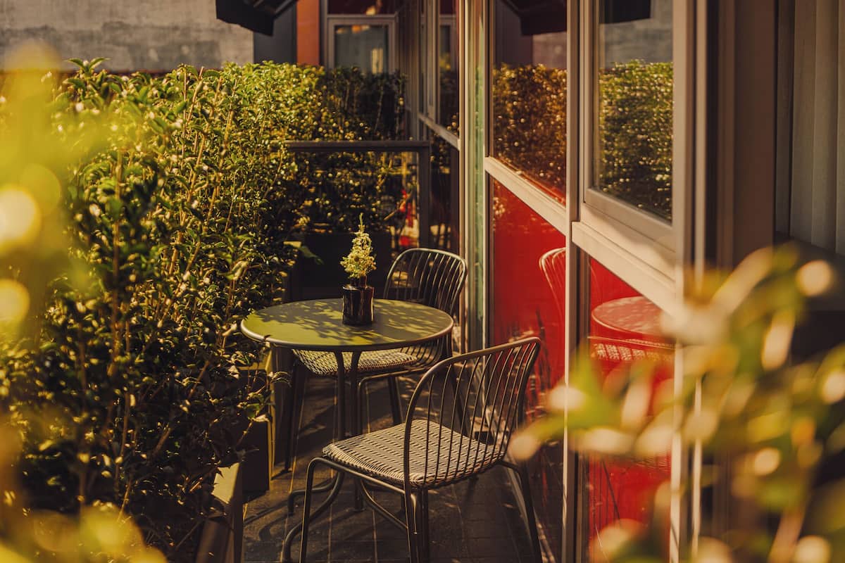 An outdoor seating area with a table and chairs, surrounded by greenery and a red wall.