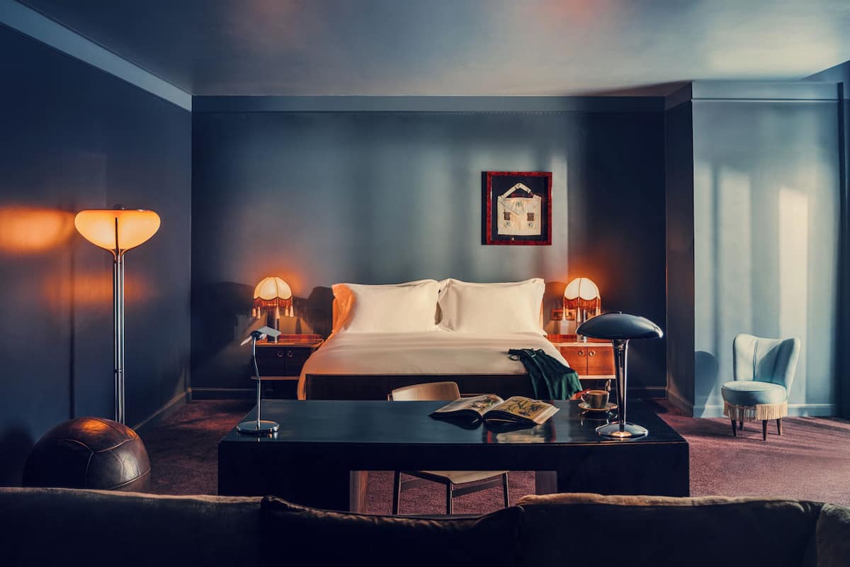 A modern hotel room with a large bed, two bedside tables, a desk with a chair, a leather armchair, and a framed picture on the wall.