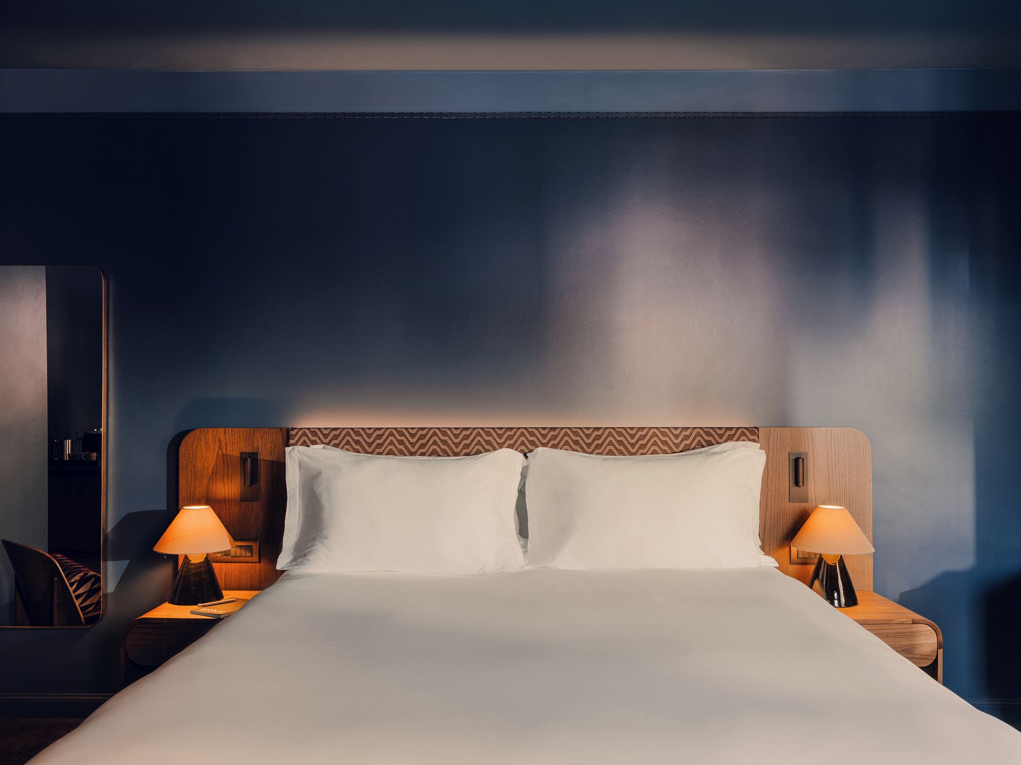 A hotel room with a large bed, two bedside tables with lamps, a wooden headboard, and a dark wall.
