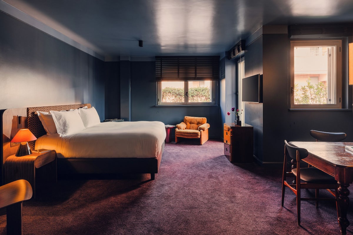 A hotel room with a bed, a desk, a chair, a sofa, and a window with blinds.