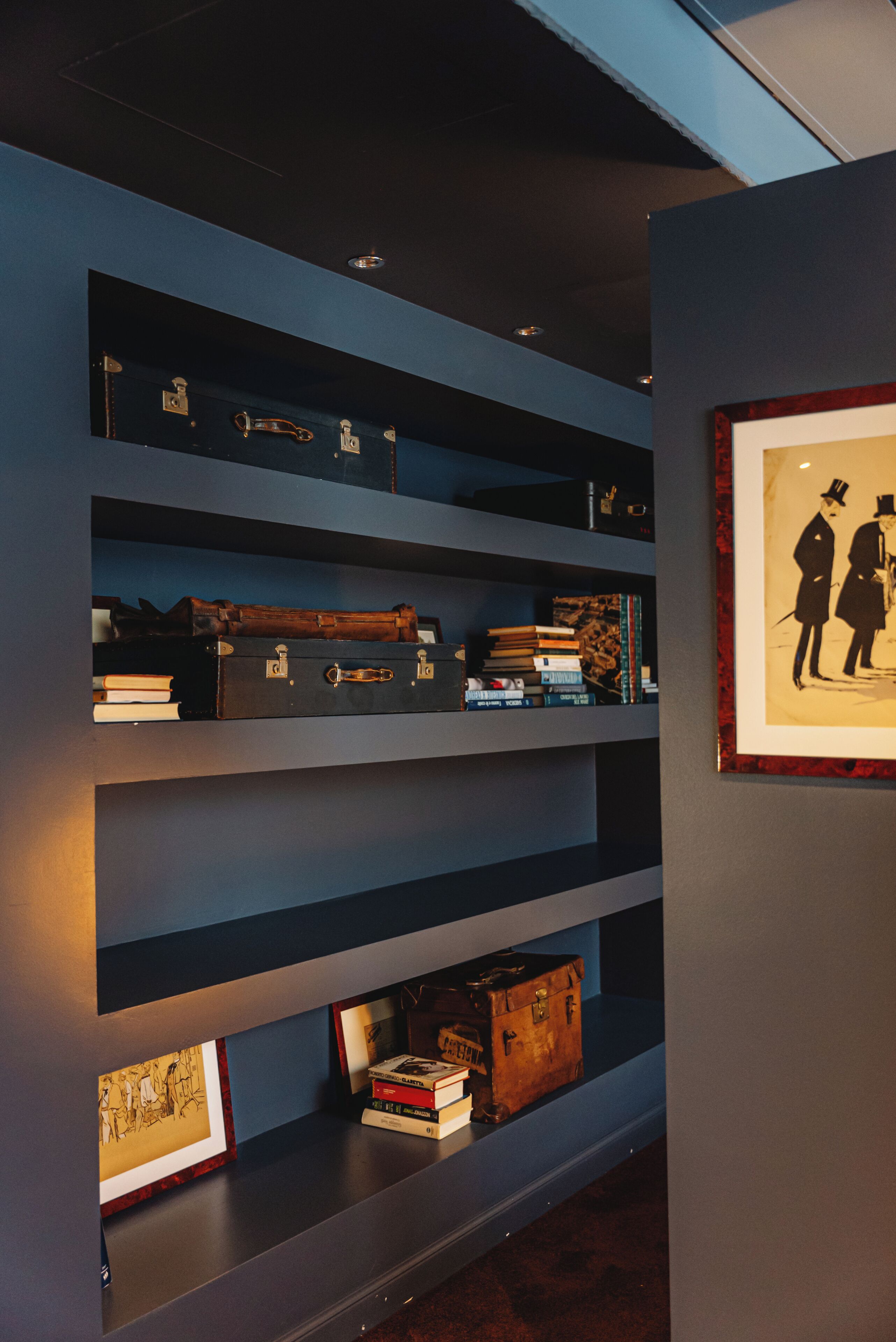 Un mur doté de plusieurs étagères où sont exposés des livres, des valises anciennes et des œuvres d’art encadrées.