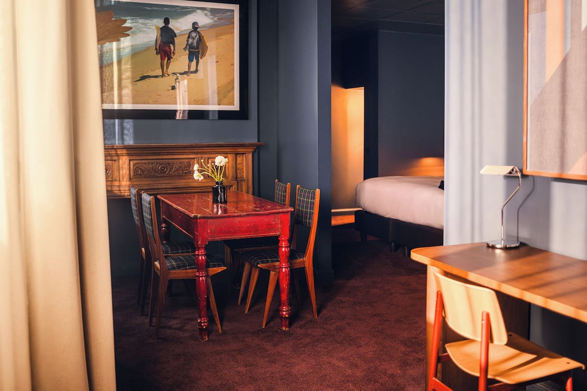 A hotel room with a wooden dining table, two chairs, a small table with a vase, a bed, a nightstand, and a painting of a beach scene.