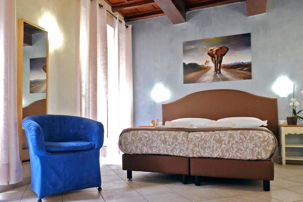 Een slaapkamer met een bed, een blauwe fauteuil, een schilderij van een olifant en een nachtkastje met een plant.