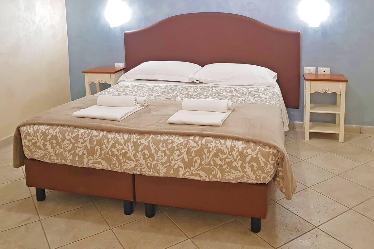Een tweepersoonsbed met een bruin hoofdeinde en een bedsprei met patroon, omringd door twee nachtkastjes met lampen, in een kamer met betegelde vloer.