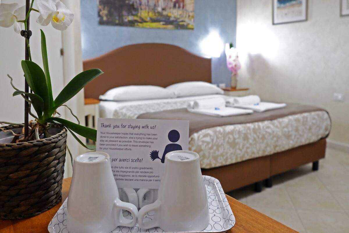 Hotelkamer met een bed, nachtkastje met een schotel witte bekertjes en een bedankkaartje, en een potorkidee.