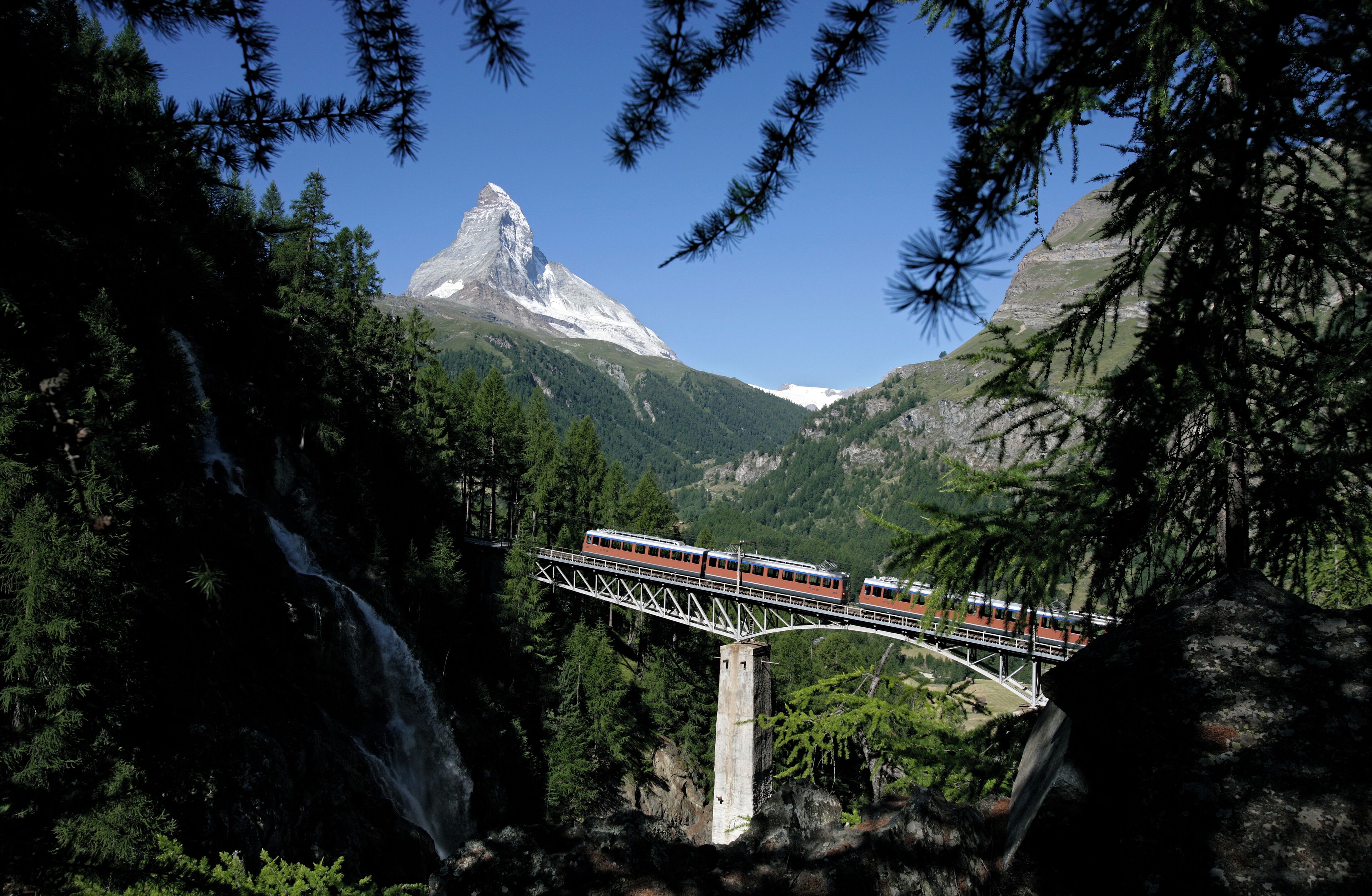 A train crossing a bridge over a waterfall in a mountainous area with a prominent peak in the background.