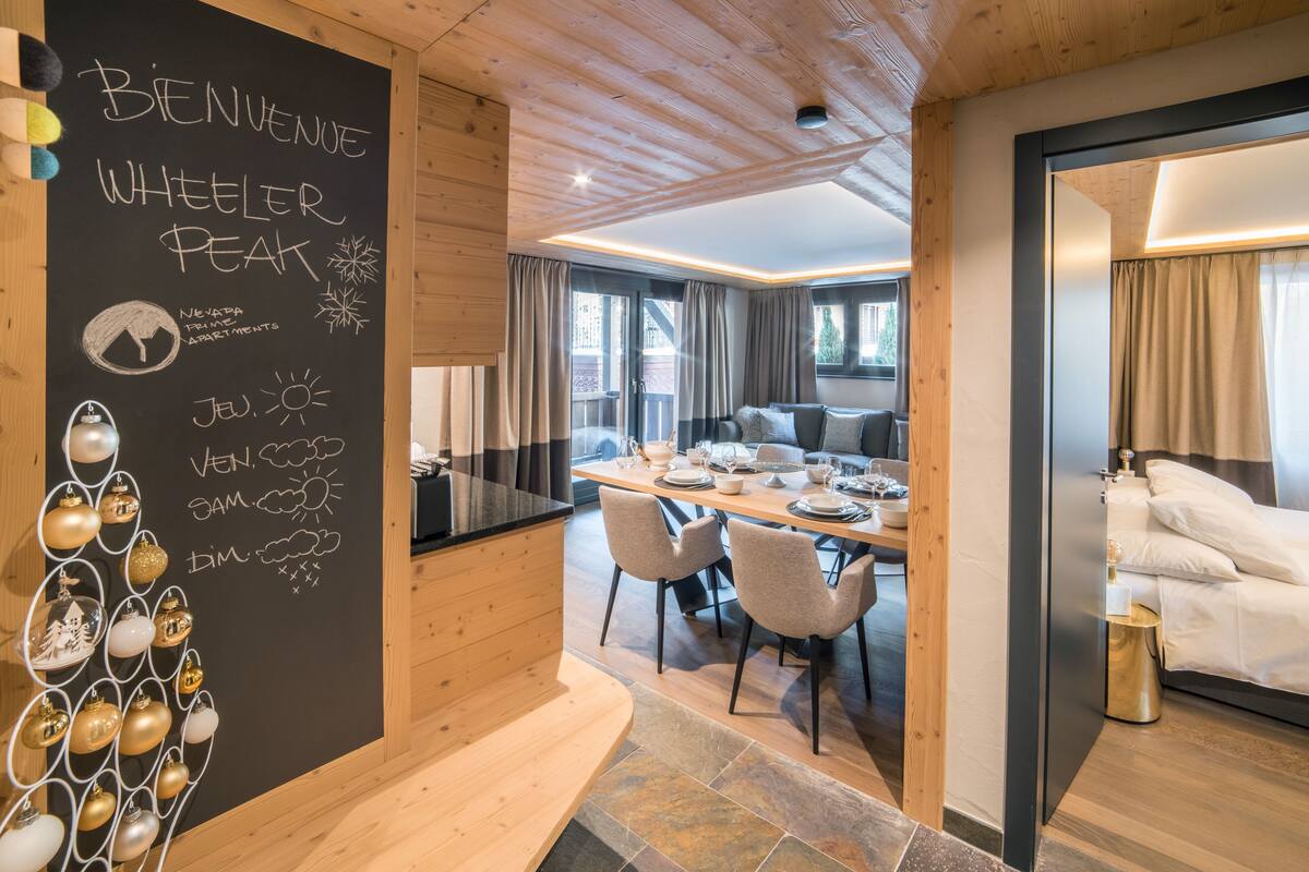 A modern hotel room with a wooden ceiling, a dining area with a table set for two, and a cozy sitting area with a sofa.