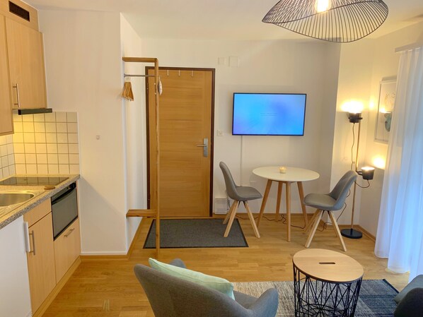 A compact living space with a kitchenette, a dining area with two chairs, a small round table, a TV mounted on the wall, and a ceiling light fixture. - Good Value Apartments by MX Zermatt (Zermatt)