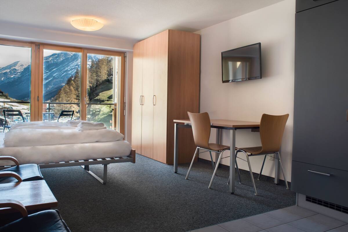 A modern hotel room with a bed, a desk, and a chair. There is a large window with a mountain view.