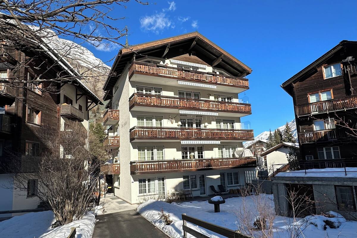 A multi-story hotel with balconies, a snow-covered ground, and a mountain backdrop.