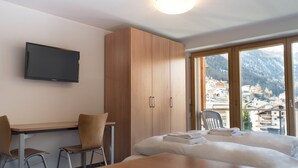 A hotel room with a bed, a desk, two chairs, a television, and a view of a mountainous town. - Good Value Apartments by MX Zermatt (Zermatt)