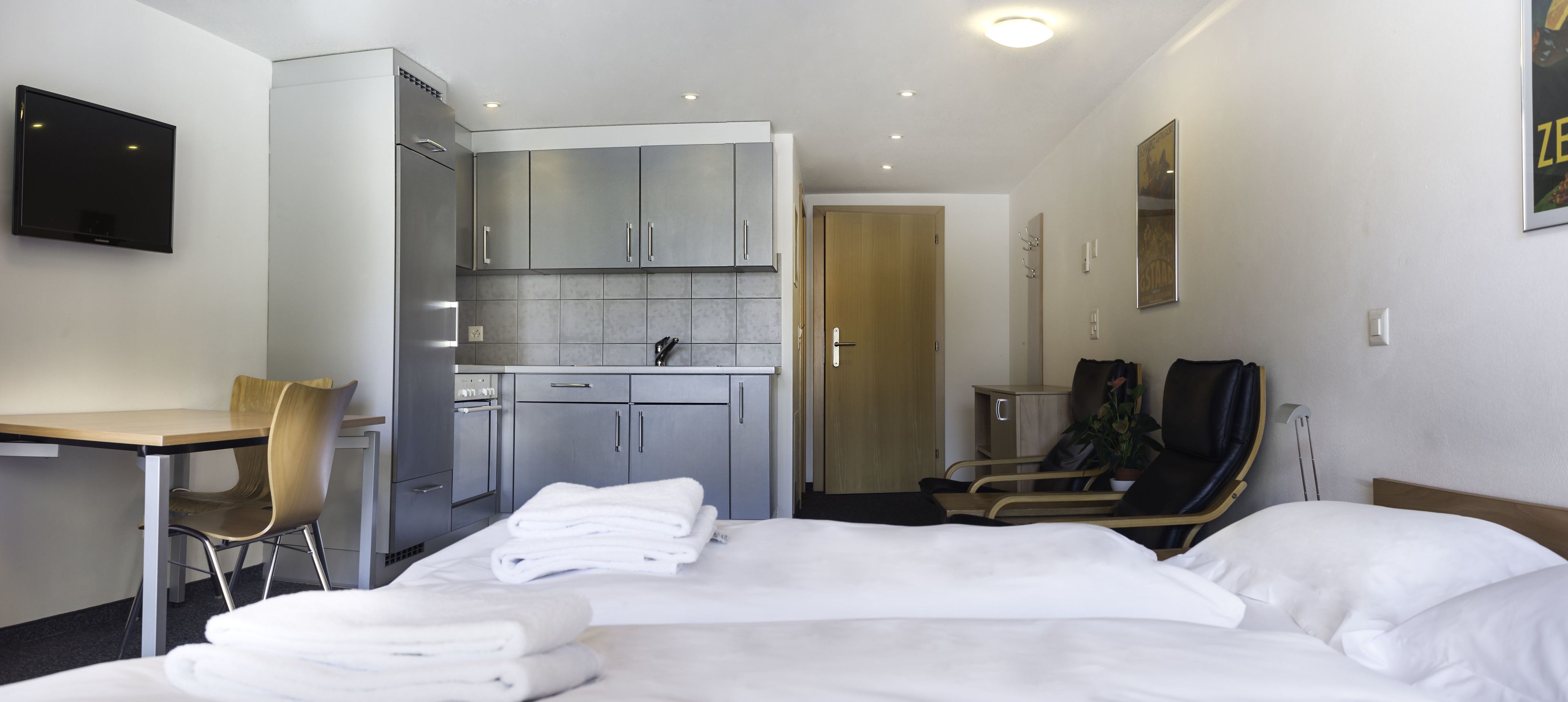 A hotel room with a bed, a desk, a chair, a kitchenette, and a bathroom.
