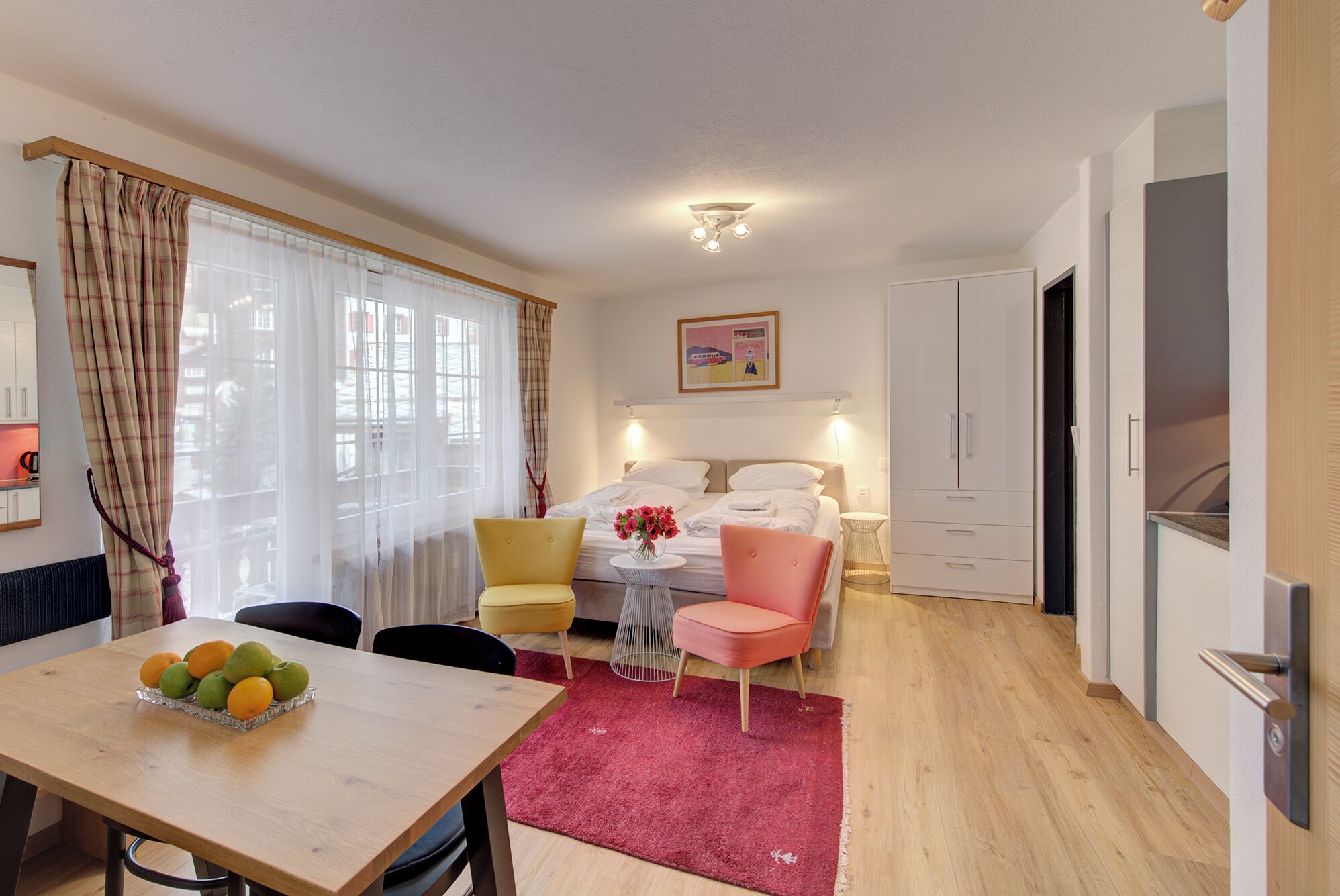 A hotel room with a bed, a dining table with fruit, a pink chair, and a red rug.