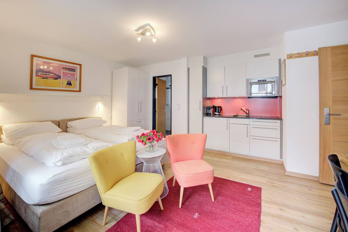 A compact hotel room with a bed, a small round table with a vase of flowers, a yellow armchair, a pink armchair, a kitchenette with a microwave, and a wooden door.