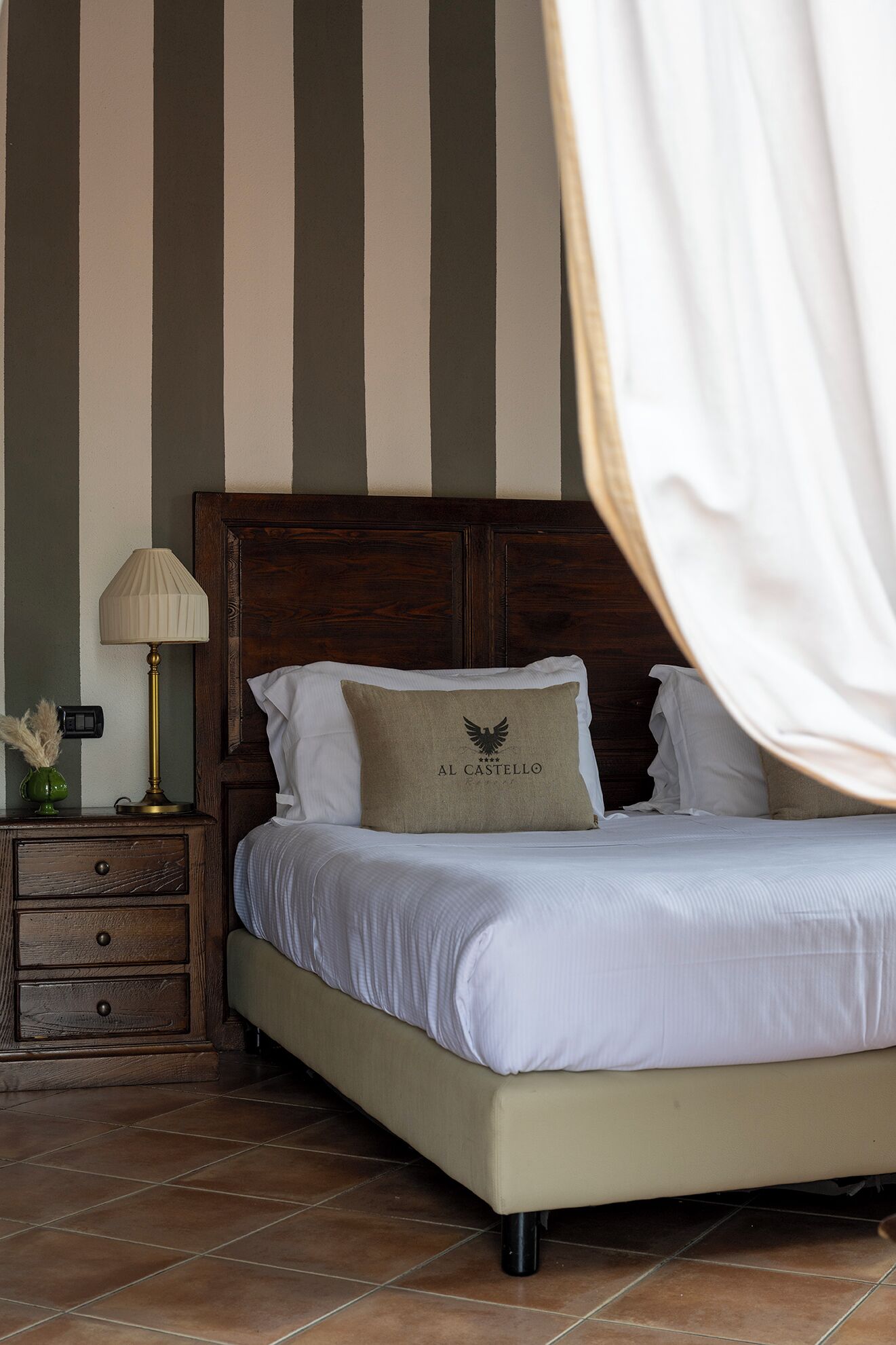 Quarto com cabeceira de madeira, cama com roupa de cama branca, mesa de cabeceira com abajur e travesseiro decorativo com logotipo.