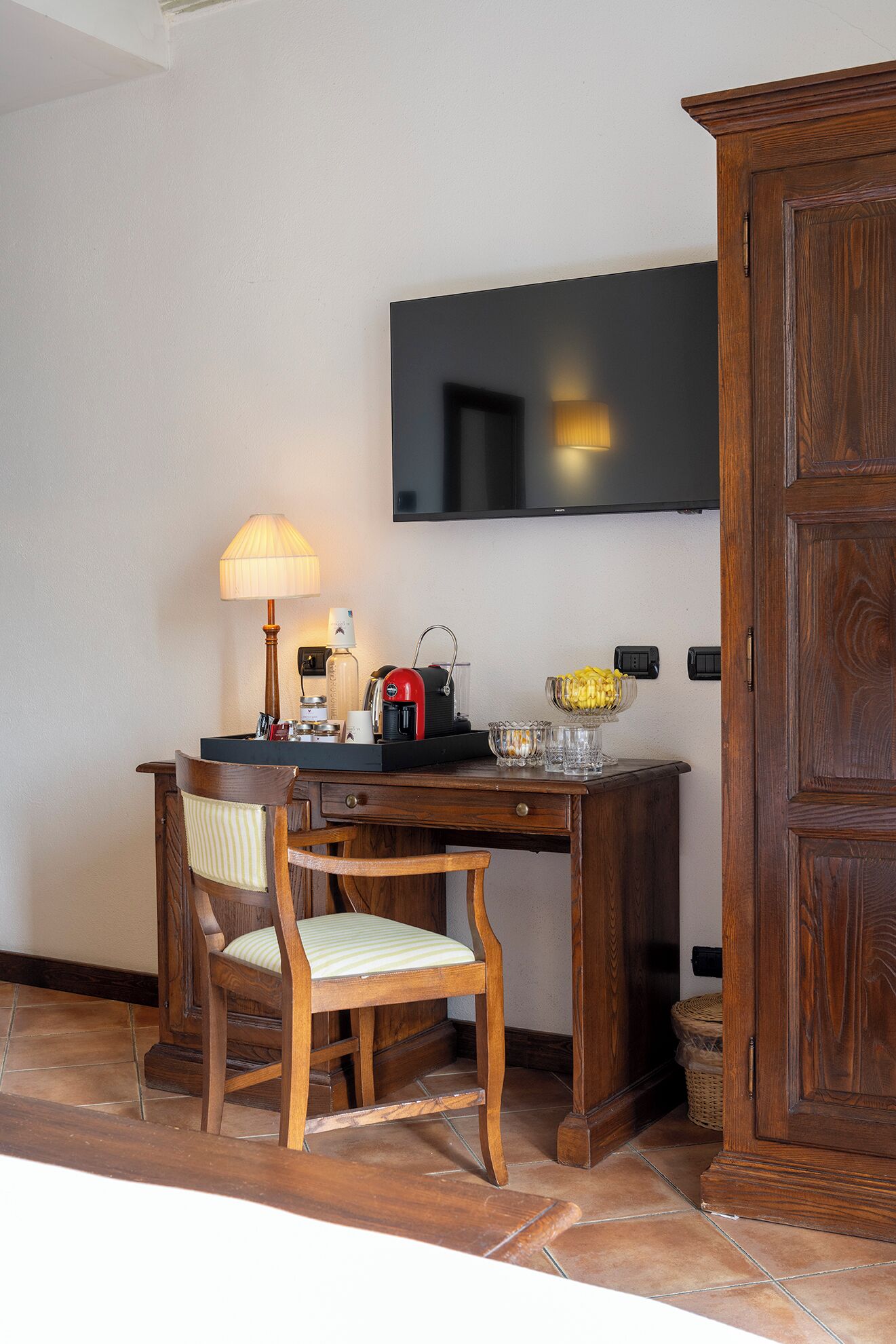 A wooden desk with a chair, a lamp, a coffee maker, and a small table with a vase of bananas.
