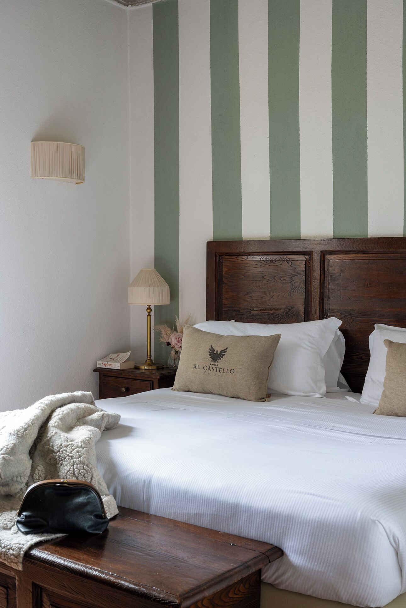 Una cama bien tendida con almohadas, una lámpara de mesita de noche y un buró de madera con lámpara.