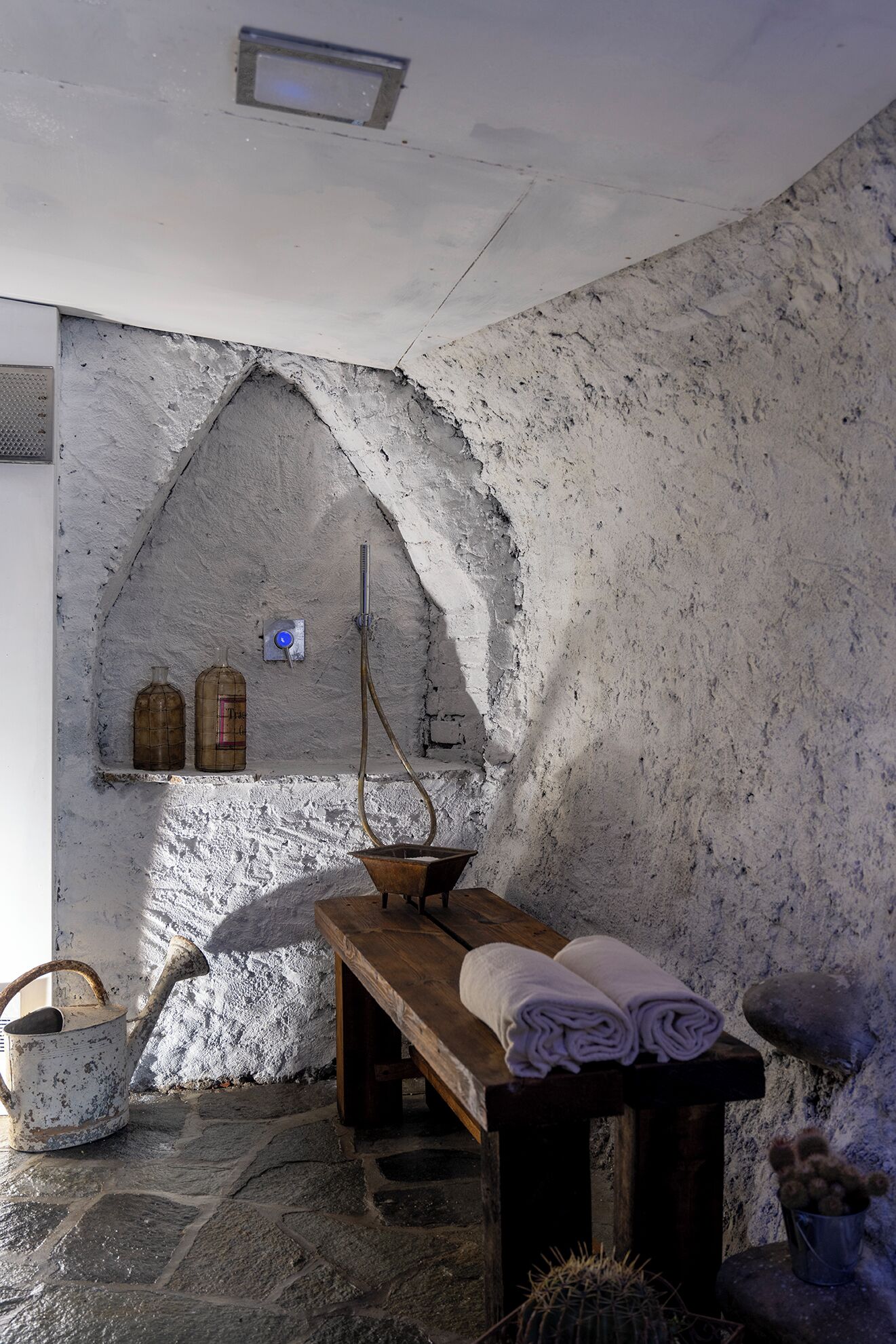 A rustic room with a stone wall, a wooden bench, a metal bucket, and a small table with towels.
