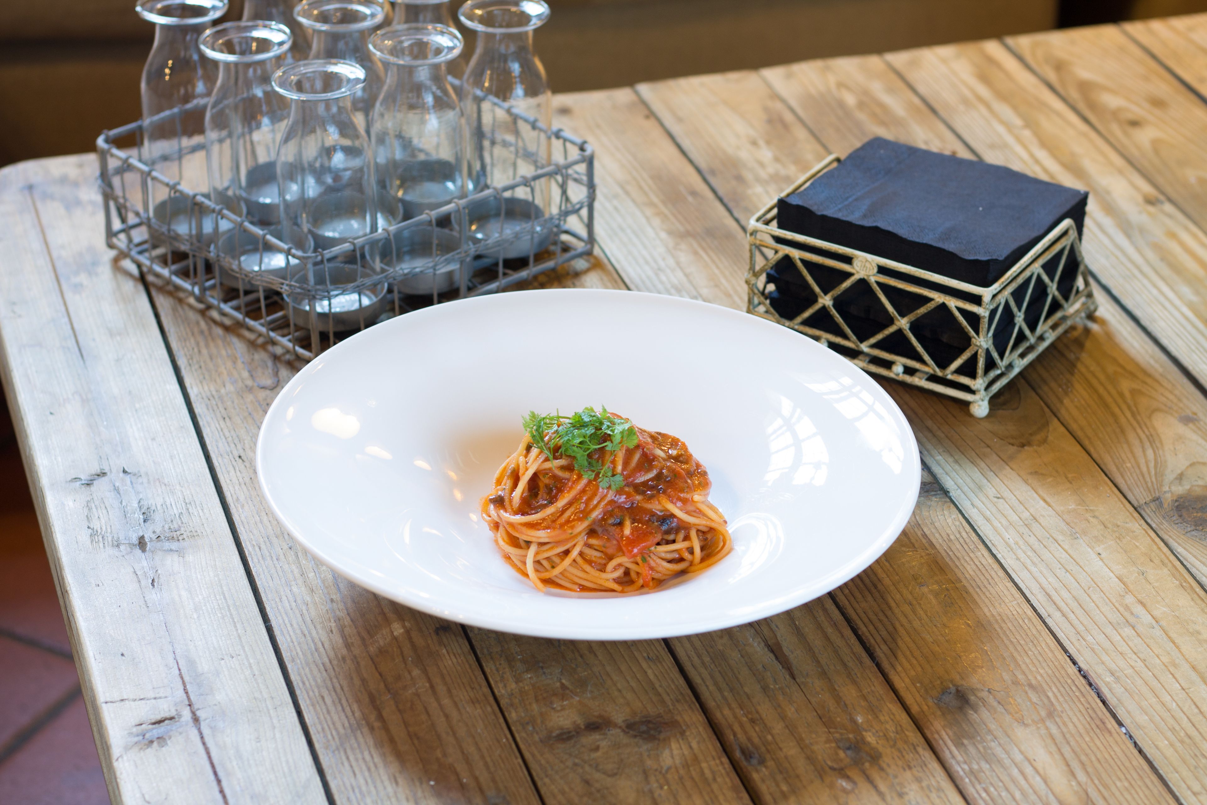 Una mesa de madera con un plato blanco de espagueti, un portavasos de vidrio y una pequeña caja negra.