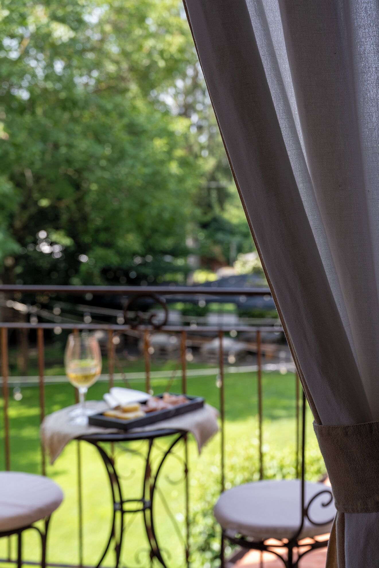 Um pátio com mesa posta para refeição, um copo de vinho e vista para um jardim verde.