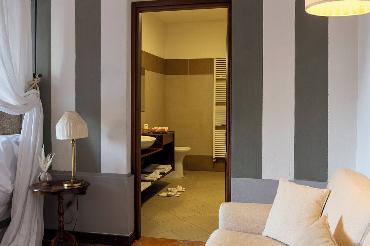 Un salón con sofá, una lámpara y una cesta tejida. Hay una puerta que da a un baño con inodoro y un perchero.