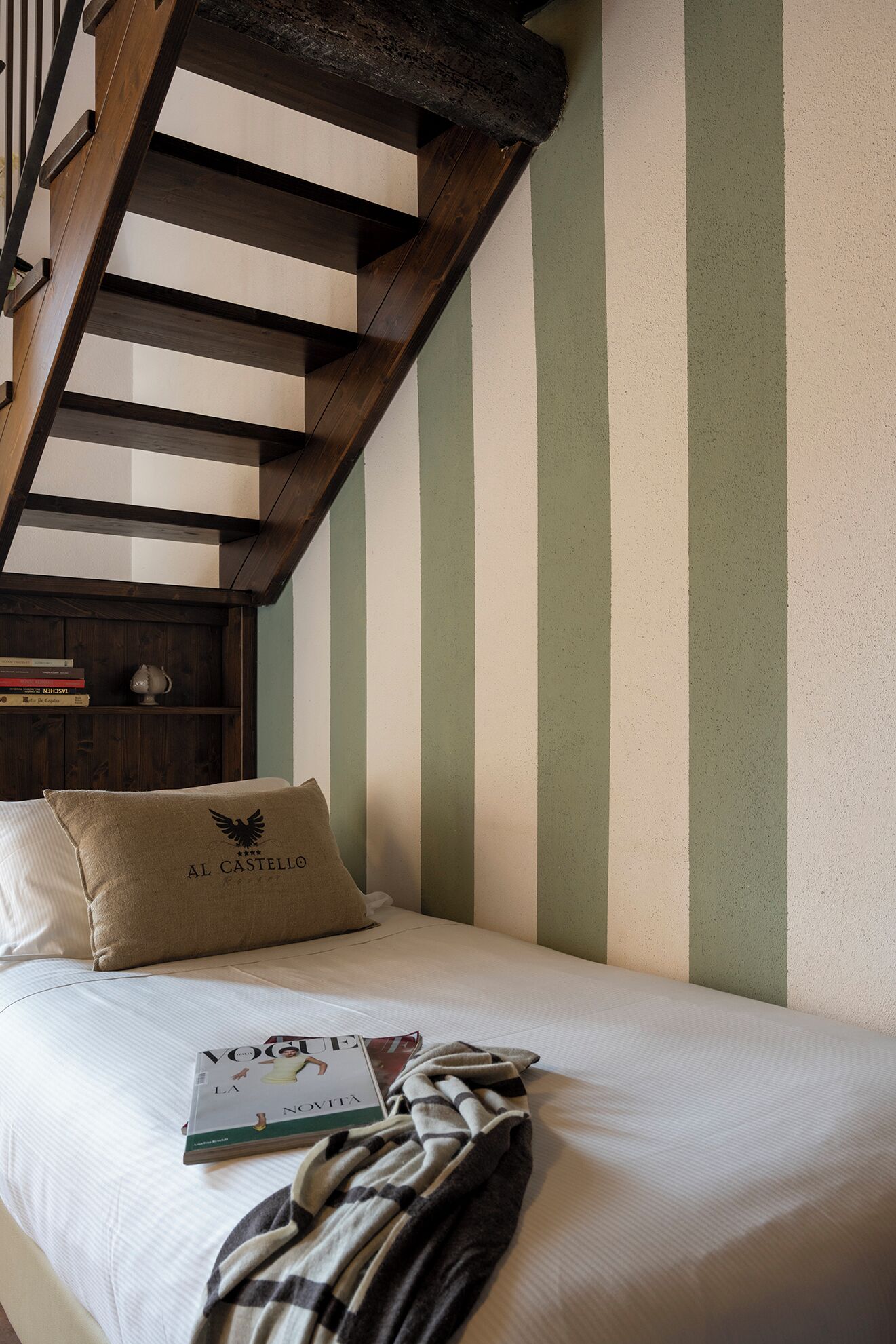 A bedroom with a wooden staircase, a bed with a pillow labeled "AL CASTELLO", and a magazine on the bed.