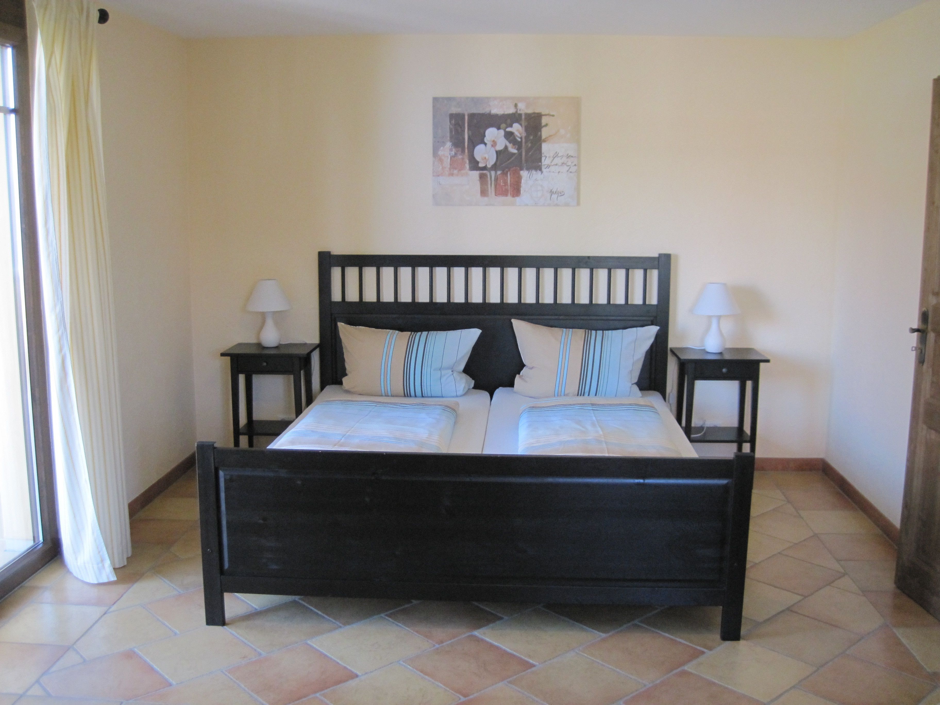 A bedroom with a dark wooden bed, two bedside tables with lamps, a window with curtains, and a wall-mounted artwork.