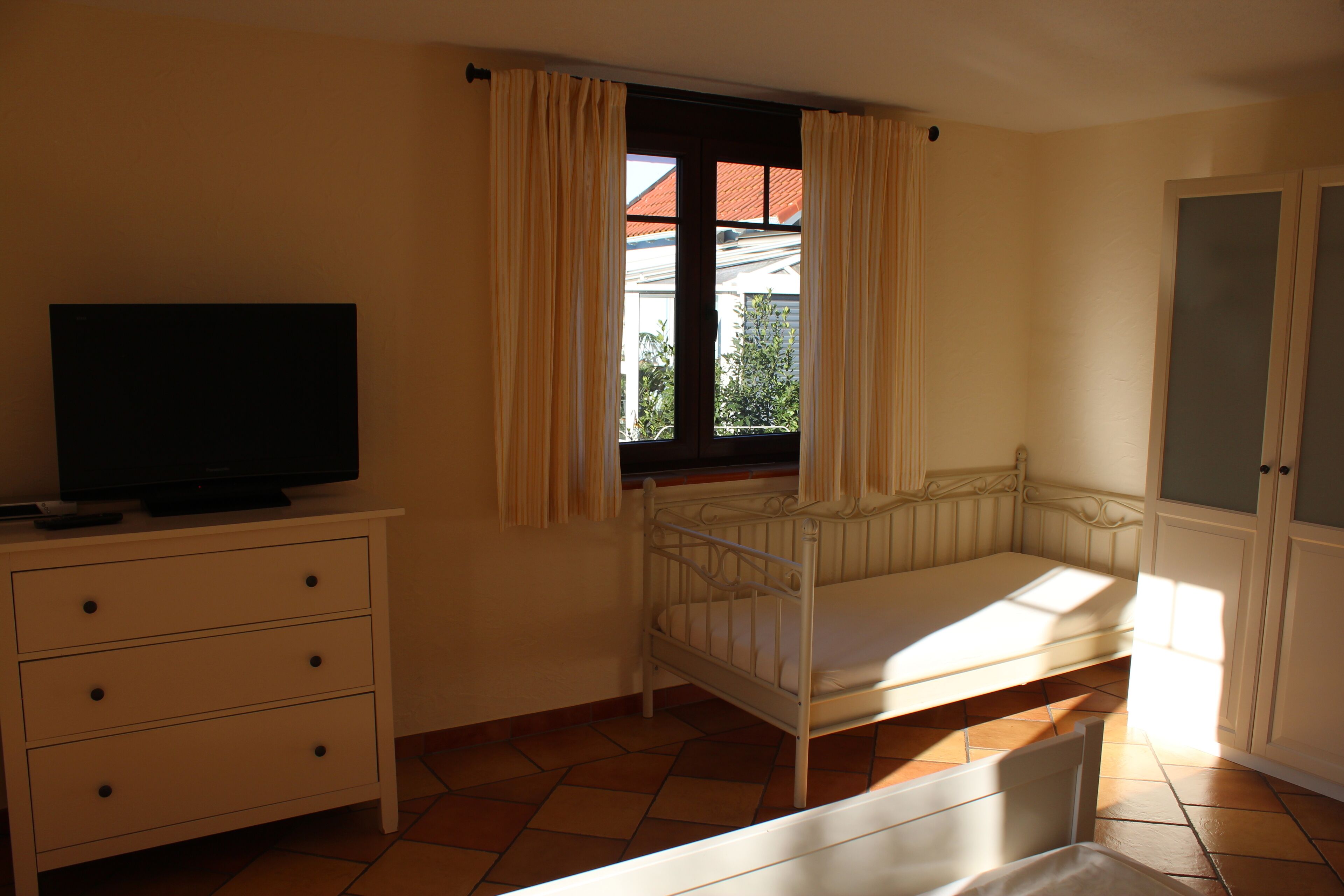 Un dormitorio con una cama individual, un televisor sobre un tocador, una ventana con cortinas y un armario.