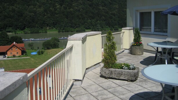 Un balcón con vistas a un lago y un bosque, muebles de exterior y un edificio con tejado de tejas.