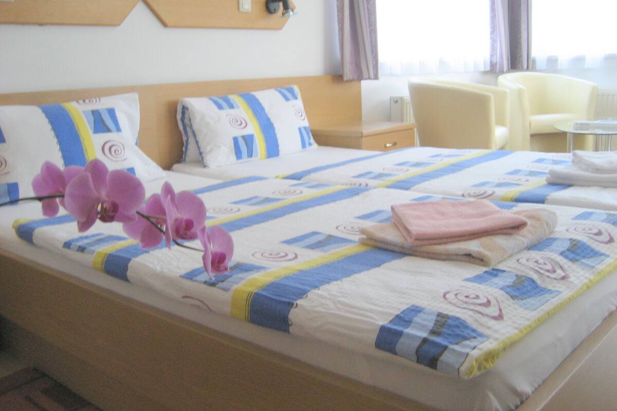 A hotel room with a bed, two chairs, a small table, and a wall-mounted panel.