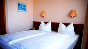 Uma cama de casal com roupa de cama branca, duas luminárias fixadas na parede e um quadro emoldurado.
