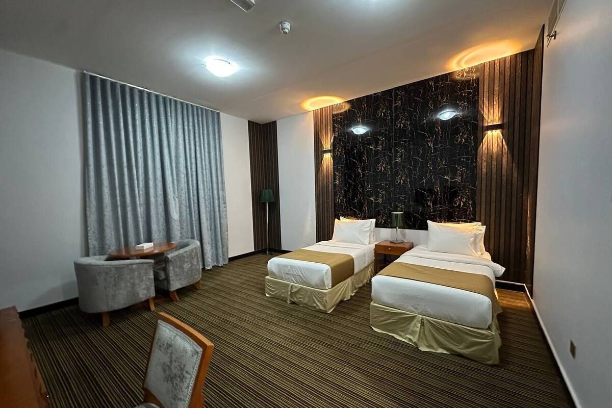 A hotel room with two single beds, a small table, a chair, and a large window with curtains.