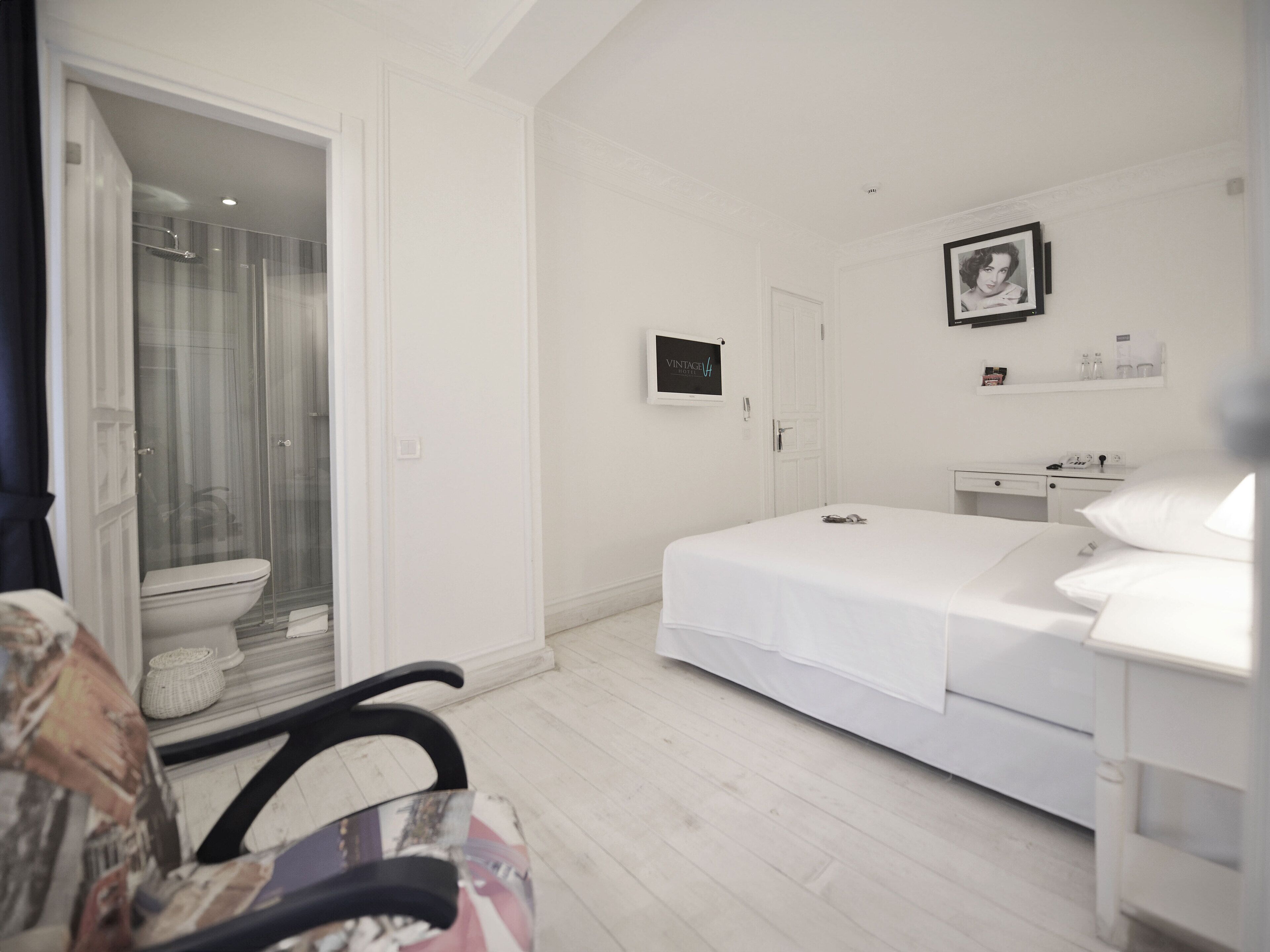 Quarto de hotel com cama, escrivaninha, cadeira, sanita e chuveiro.