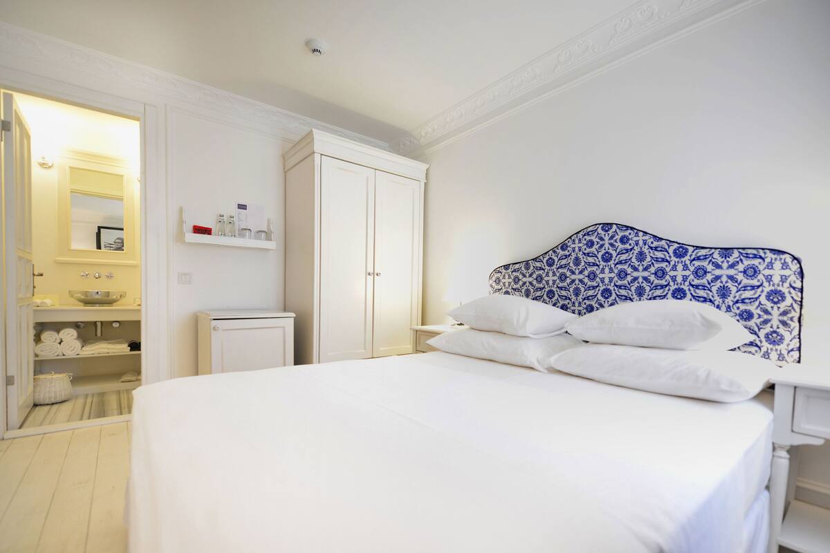 Une chambre à coucher avec un grand lit, une armoire, une table de chevet et une salle de bain visible à travers une porte ouverte.