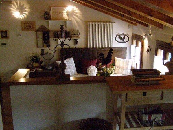 Una habitación acogedora con techo de madera, un radiador, una cama con adorno de gallo, una estantería con libros y un jarrón de flores, y un cartel que dice 'Bienvenue'.